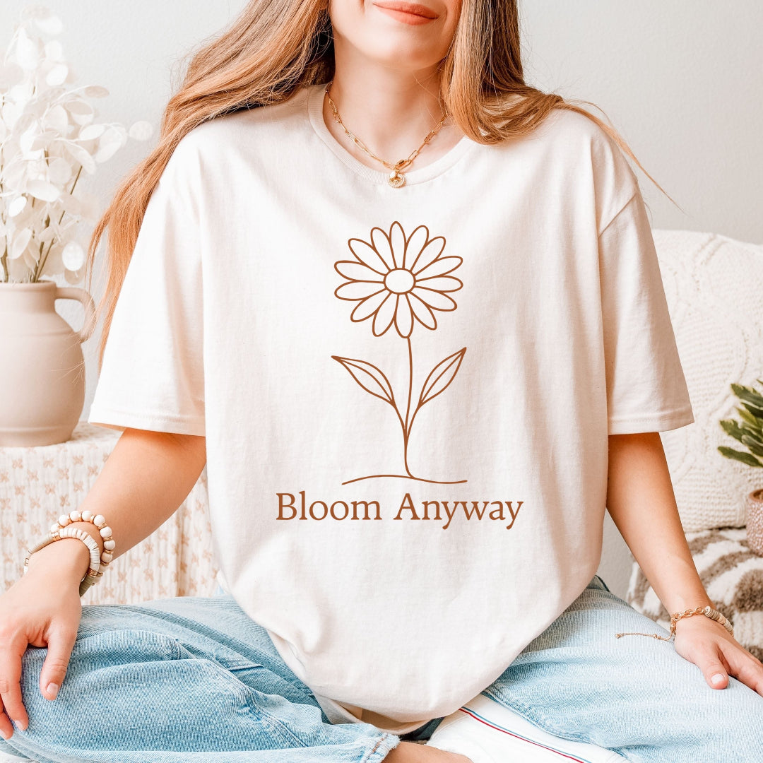 A person wearing a natural colour t-shirt with a terracotta line-art flower graphic and 'Bloom Anyway' text underneath.