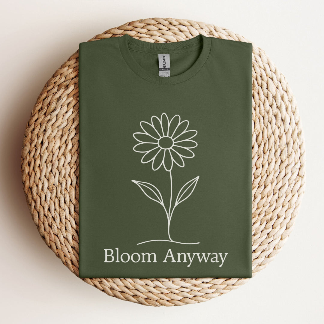 A folded military green t-shirt with a creamy off-white line-art flower graphic and 'Bloom Anyway' text underneath.