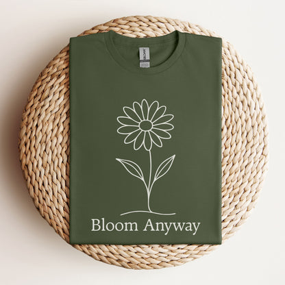 A folded military green t-shirt with a creamy off-white line-art flower graphic and 'Bloom Anyway' text underneath.