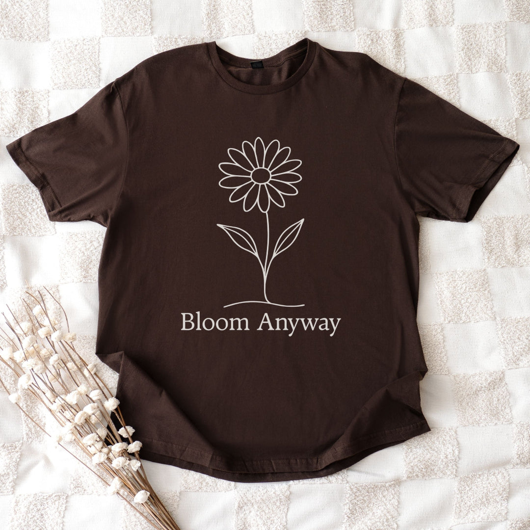 A flat lay dark chocolate colour t-shirt with a creamy off-white line-art flower graphic and 'Bloom Anyway' text underneath.